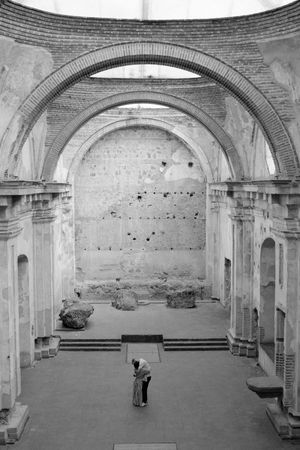 Black and white photo of a lone person standing inside ancient ruined stone arched building with exposed brick walls.