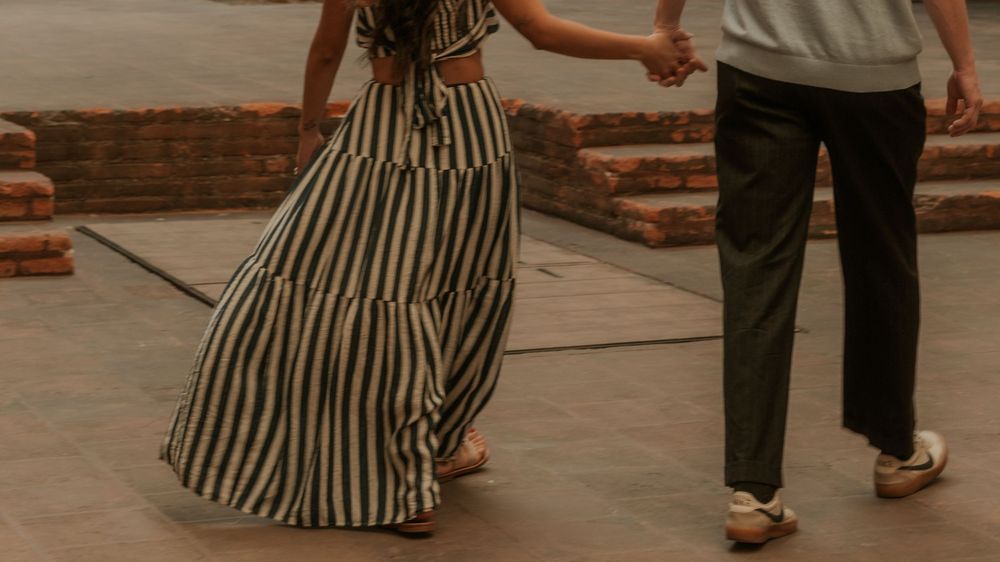 A couple holds hands while walking together, the woman wearing a flowing striped maxi dress and the man in black trousers.