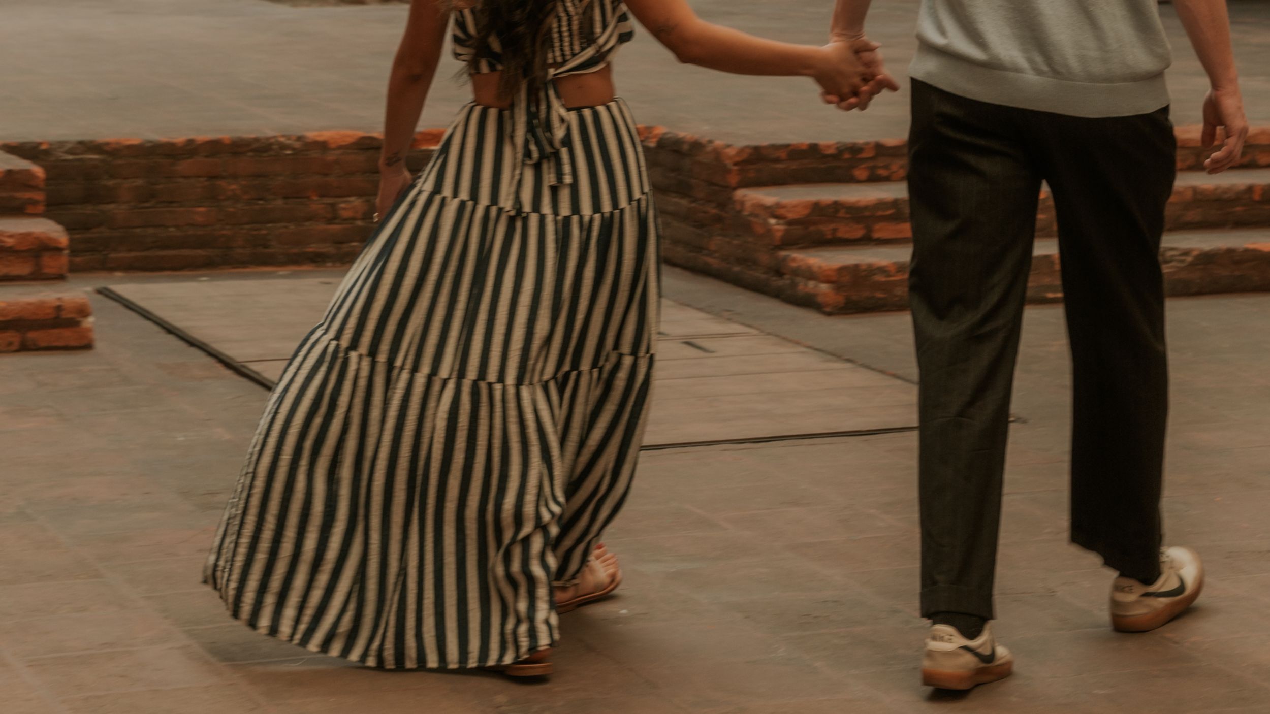 A couple holds hands while walking together, the woman wearing a flowing striped maxi dress and the man in black trousers.
