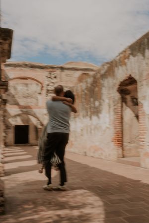 Couple embracing romantically among ancient ruins with weathered brick walls and arched doorways under a cloudy sky.