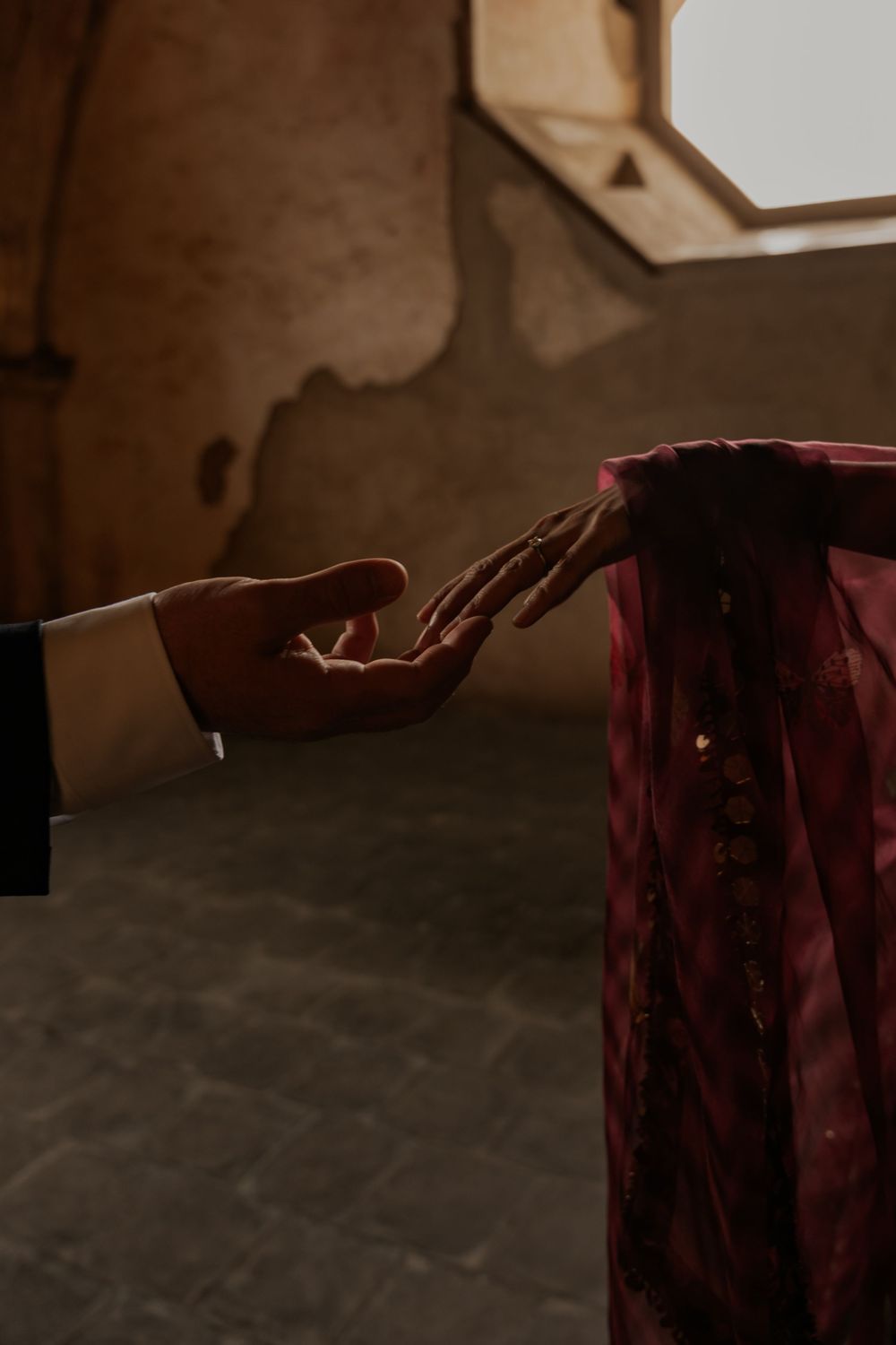 A groom in a suit gently holds the hand of a bride wearing a red embroidered dress in a rustic setting.