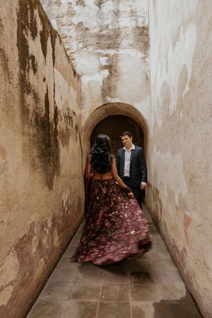 Couple in a narrow stone corridor, woman in floral burgundy gown twirling toward man in navy suit near arched doorway.