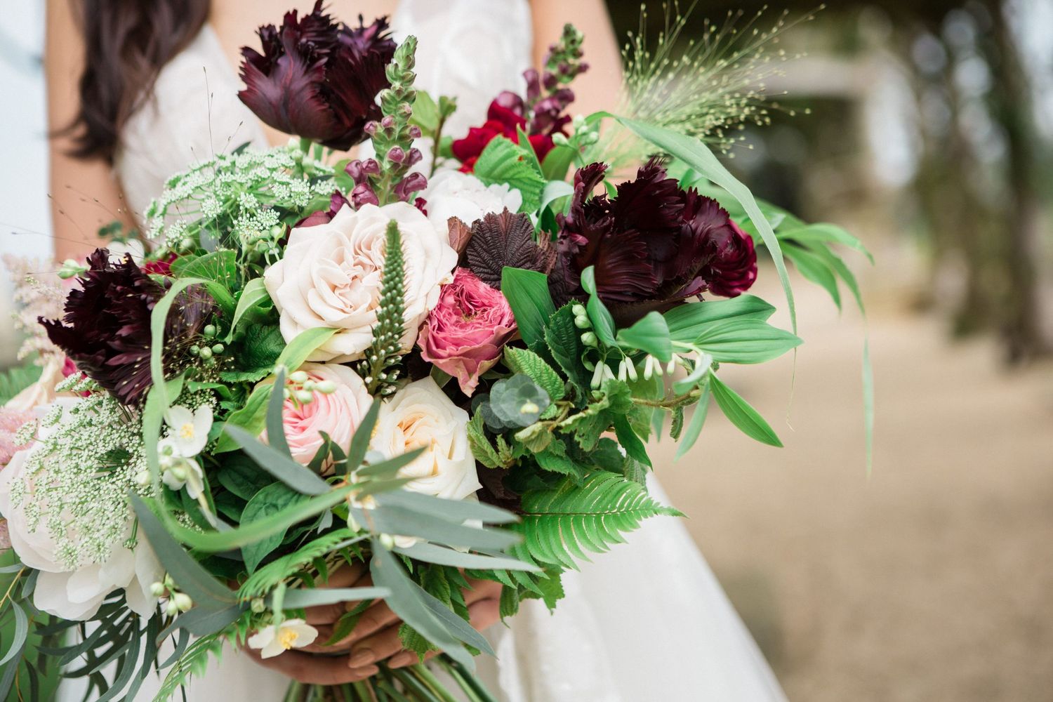 Bride holding lush wedding bouquet with dark burgundy peonies, blush roses, and greenery.