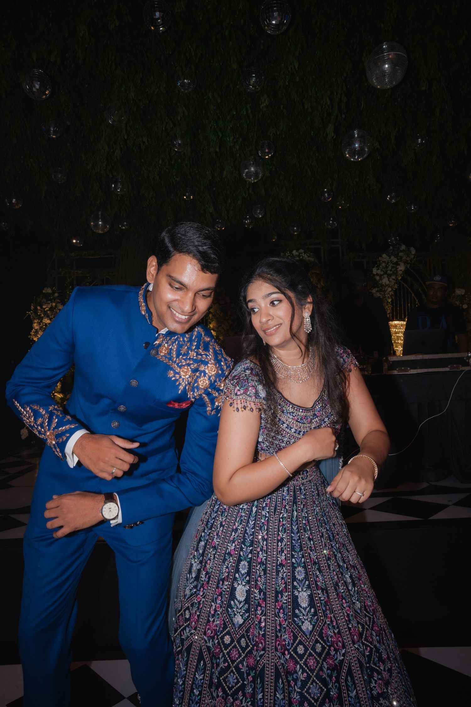 Couple dancing joyfully at Indian wedding, man in blue embroidered suit, woman in grey embroidered lehenga.
