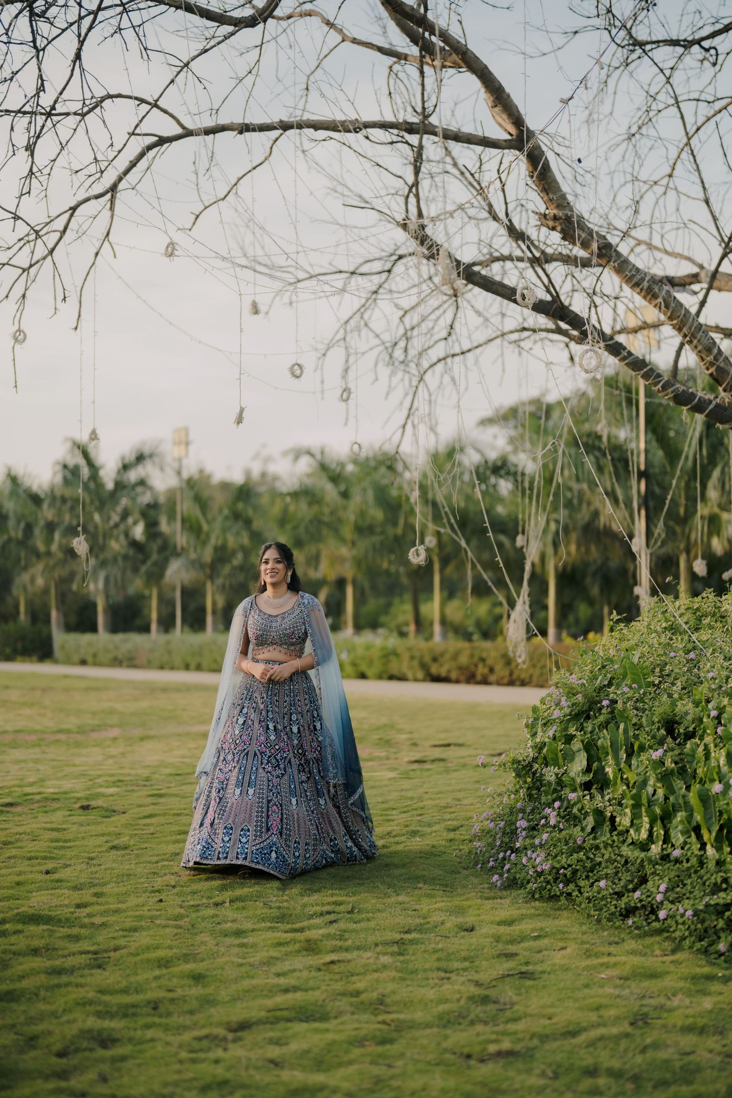 Woman in blue embroidered lehenga standing on green lawn under bare tree with hanging decorations.