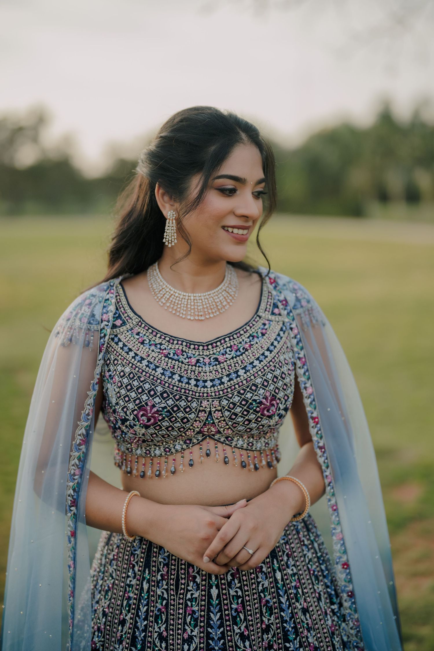 Indian bride in embroidered grey lehenga with pearl necklace and earrings, smiling outdoors at dusk.