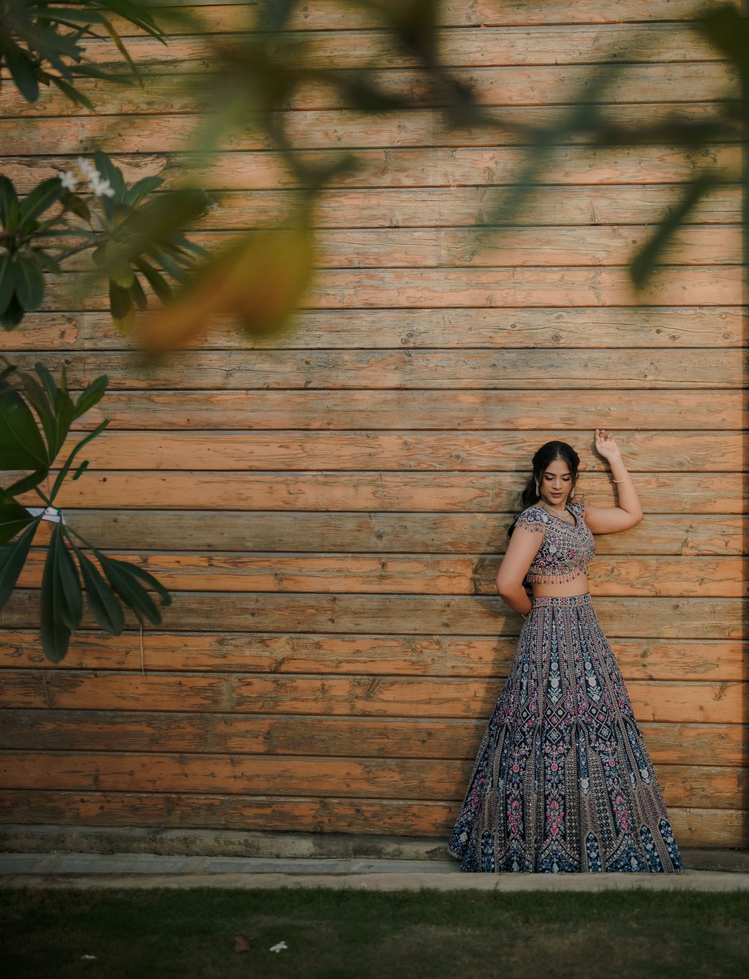 Woman in embroidered blue lehenga posing gracefully against wooden wall backdrop in golden hour light.