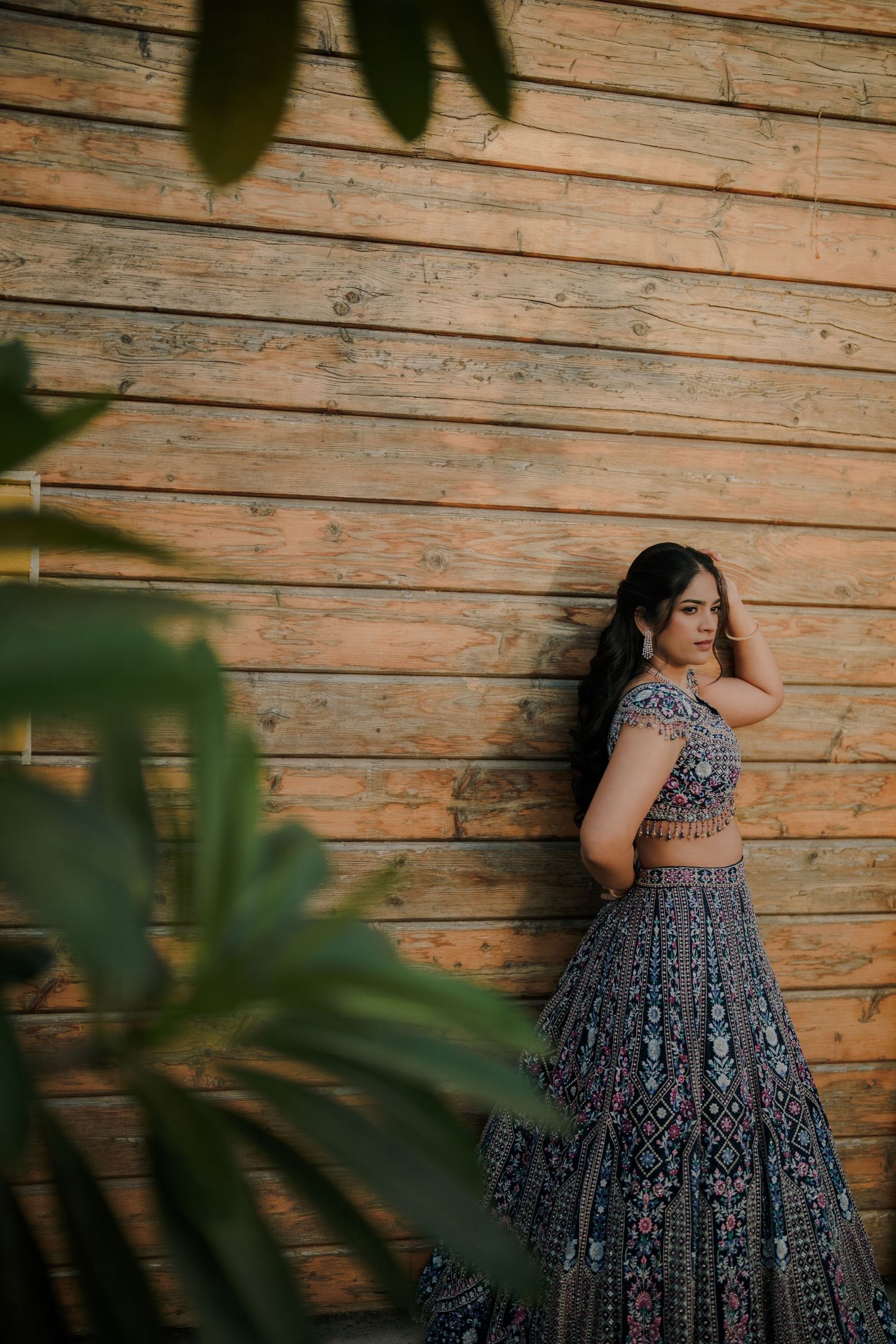 Woman in embroidered blue lehenga poses gracefully against a warm wooden wall with lush green plants in foreground.