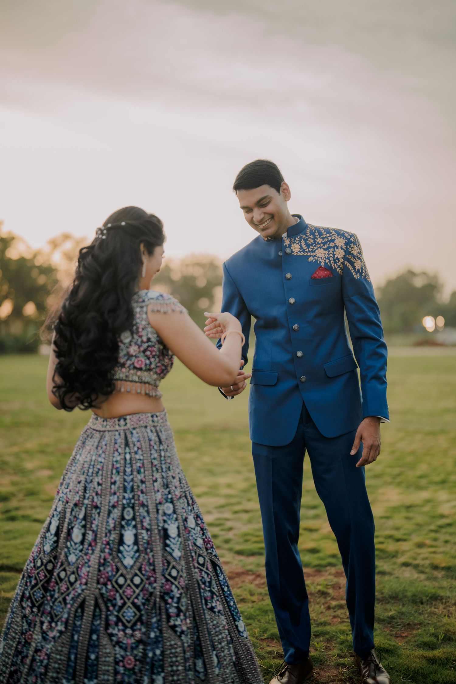 Indian couple in coordinating blue outfits holding hands on green lawn at sunset.