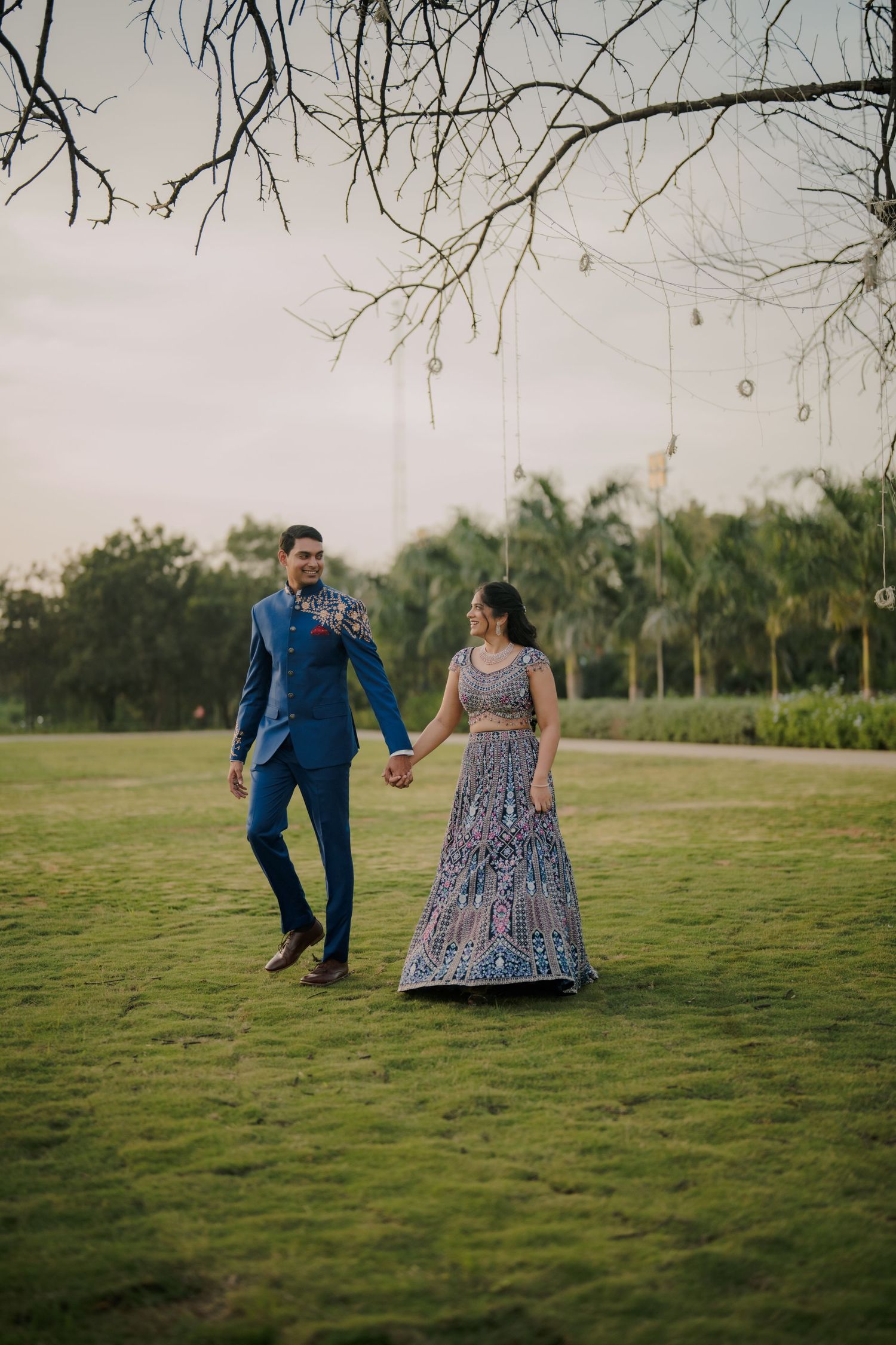 Couple walking hand in hand on green lawn, man in blue suit, woman in embroidered lehenga, with trees in background.