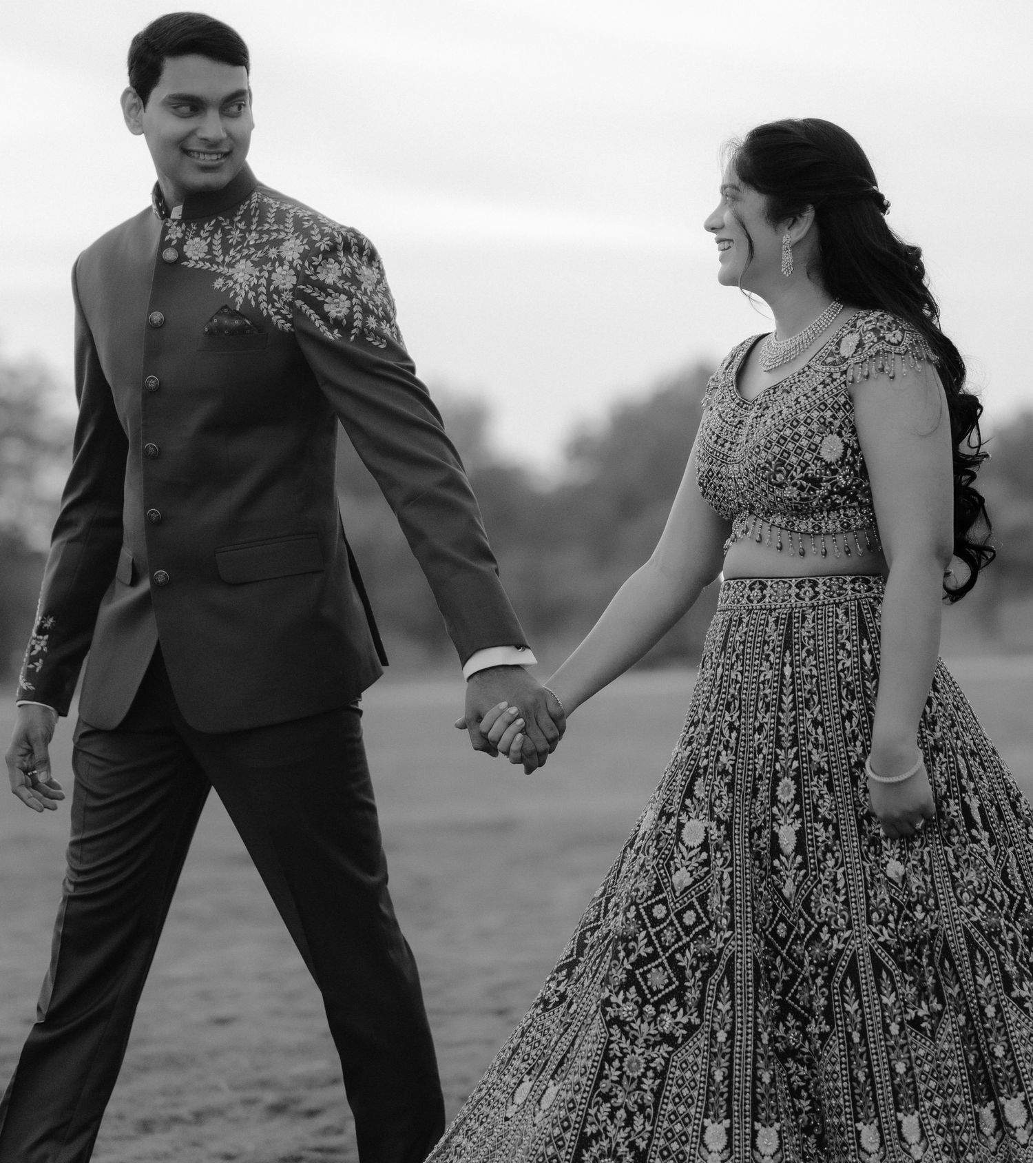 Black and white photo of an Indian couple in traditional attire holding hands outdoors during a wedding celebration.