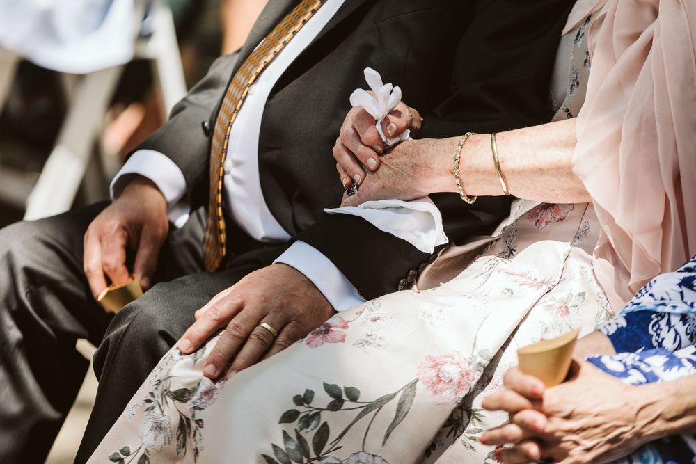 Close-up of wedding guests holding hands during a ceremony, wearing elegant attire with floral dress details.