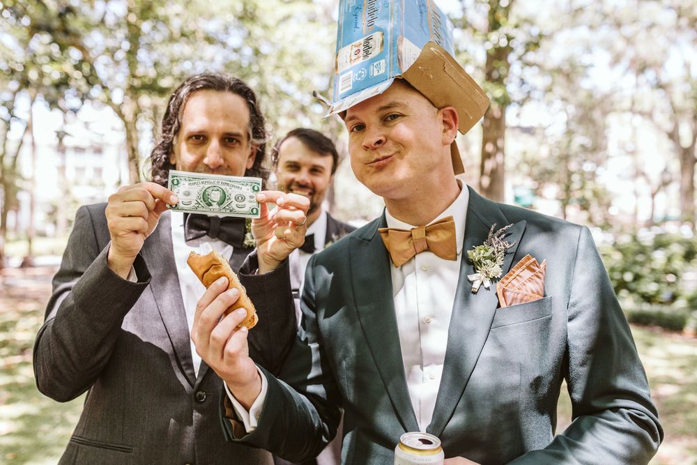 Two grooms in suits celebrate at wedding, one holding dollar bill, other wearing paper hat, with bow ties outdoors.