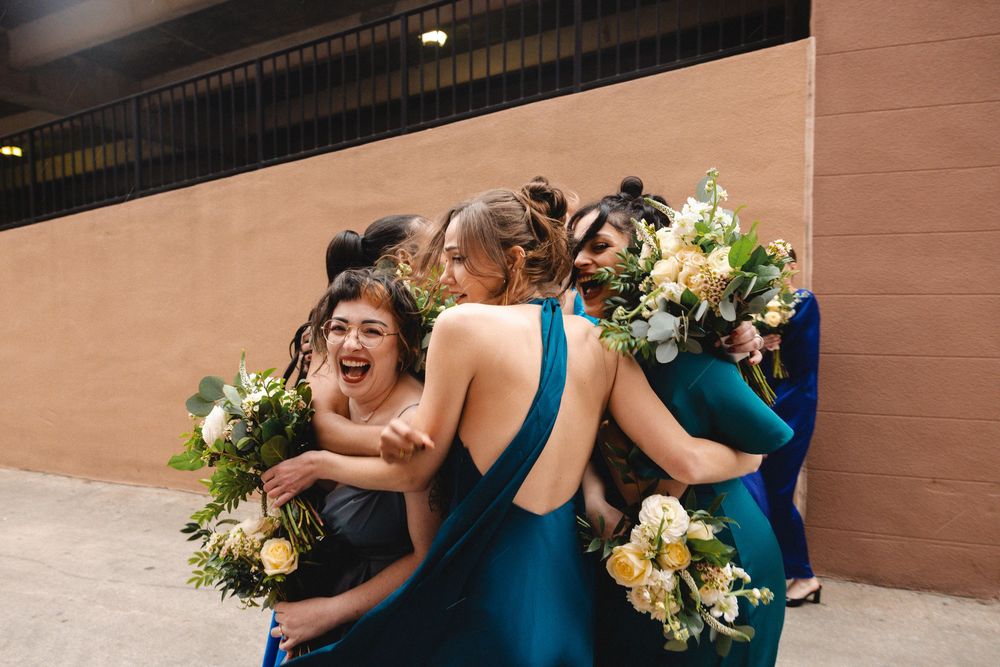 Bridesmaids in teal and navy dresses hugging and laughing together while holding white floral bouquets outdoors.