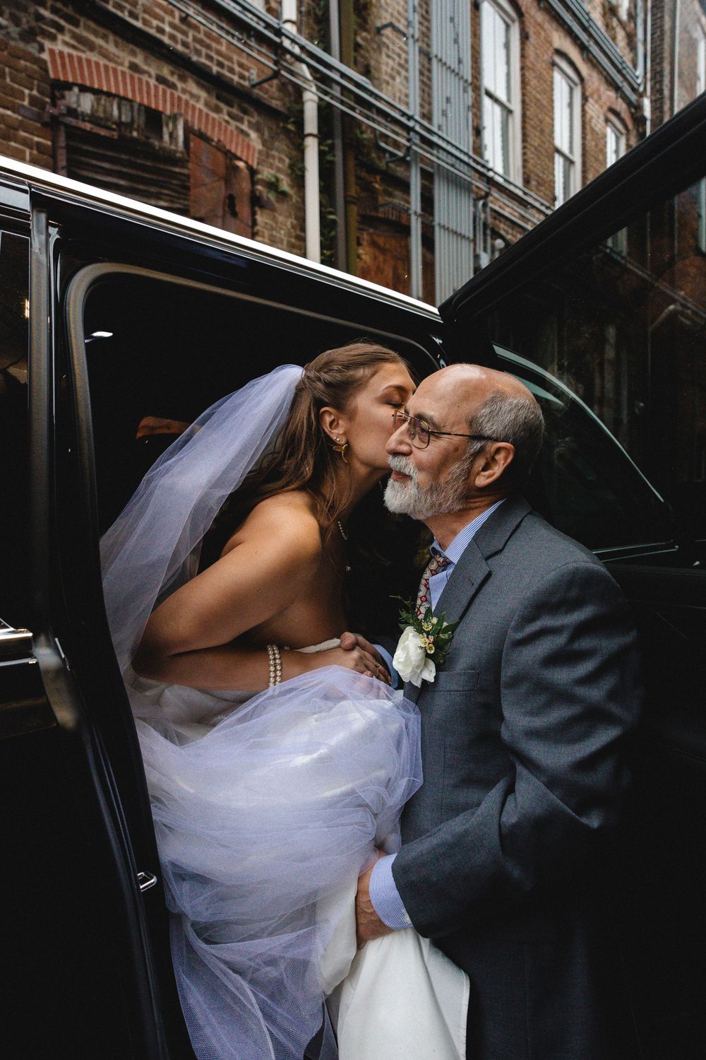 Bride in white gown and veil shares a tender kiss with older man in gray suit beside black vehicle.