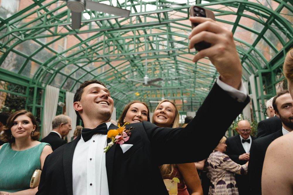 Groom in tuxedo takes selfie with smiling guests inside elegant green glass atrium at wedding reception.