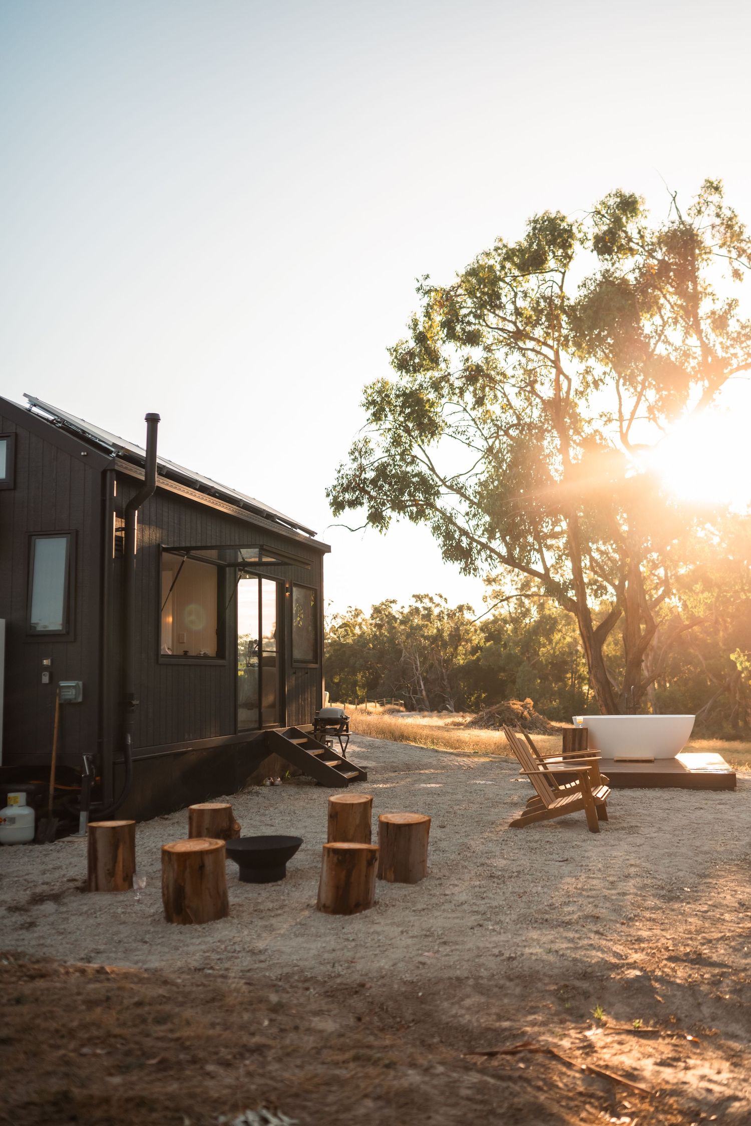 Rustic tiny house at sunset with outdoor seating made from tree stumps and a freestanding bathtub. Golden sunlight filters through a large