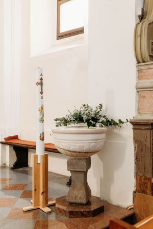 Stone baptismal font adorned with white flowers beside a tall Easter candle on wooden stand in a church.