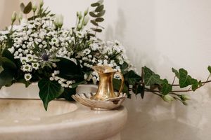 Gold vintage candlestick holder beside white floral arrangement with ivy on marble surface.