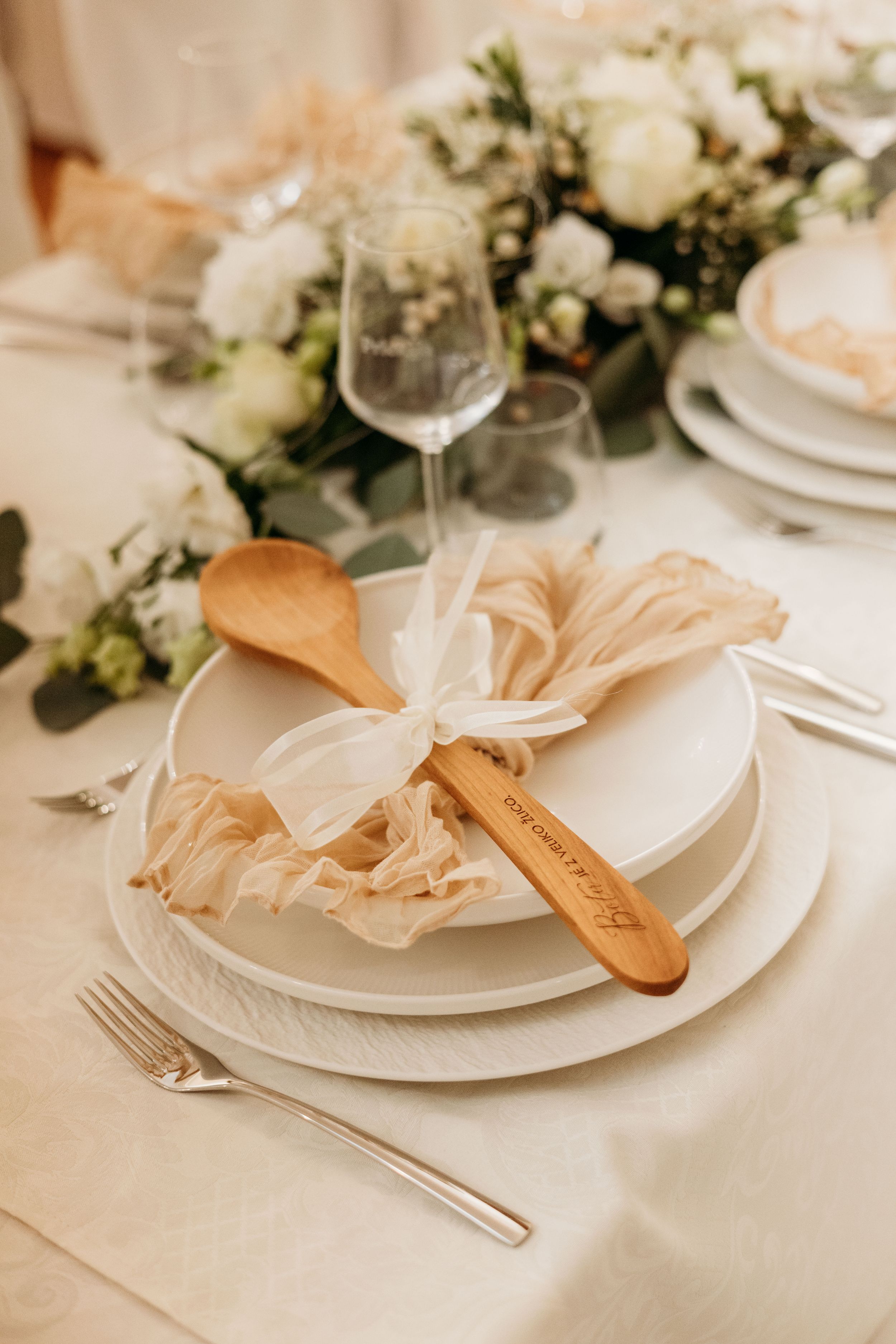 Elegant wedding place setting with wooden spoon, white ribbon bow, stacked plates, crystal glassware, and white floral centerpiece.
