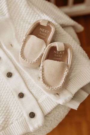 Cream Next Baby moccasin loafers placed on a white knit button-up baby cardigan sweater.