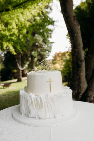 Two-tier white wedding cake with ruffled fondant, gold cross, and decorative topper displayed outdoors.