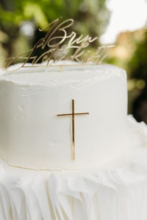 Two-tier white wedding cake with gold cross decoration and personalized script cake topper on outdoor background.