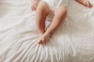 Chubby baby legs and feet resting on a soft white bed, wearing a white onesie.