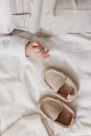 Newborn baby hand next to beige Next Baby moccasin loafers on white bedding.