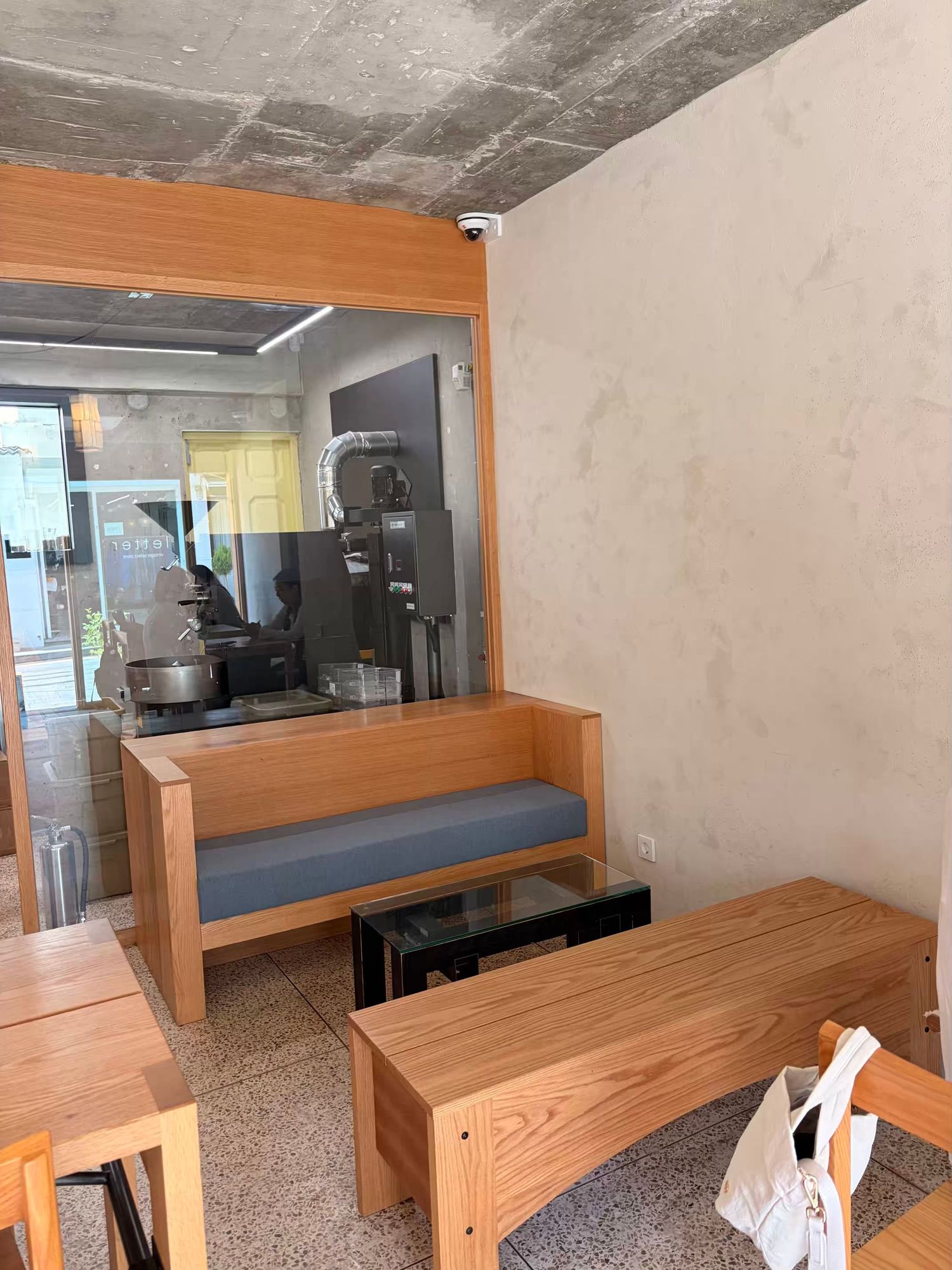 Minimalist cafe interior with light wood furniture, blue cushioned bench, glass partition revealing kitchen equipment.