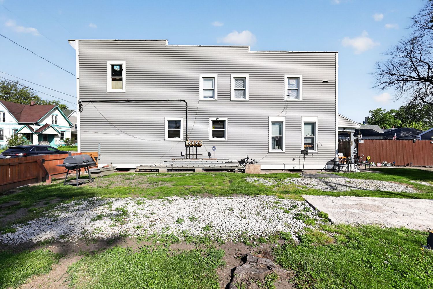 Rear exterior of a two-story gray duplex with a large backyard, patio area, and wooden fence on a sunny day.
