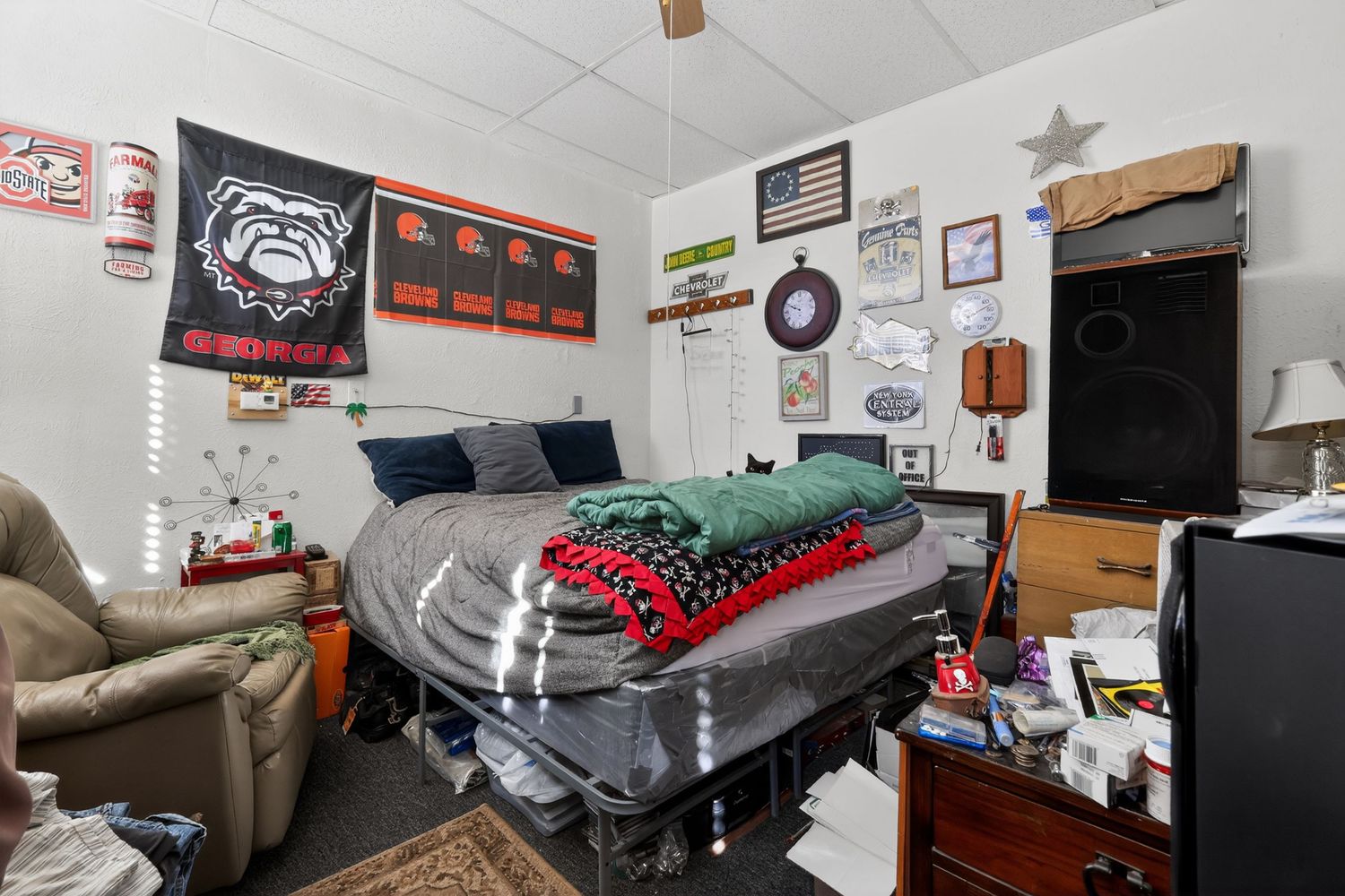College dorm room with UGA Bulldogs banner, messy bed with red blanket, recliner chair, and American flag wall decor.
