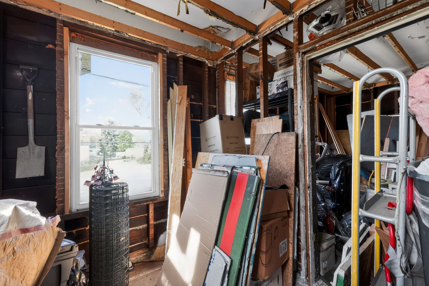 Cluttered storage room with wood paneling, a window, leaning boards, wire fencing, and tools on shelves.
