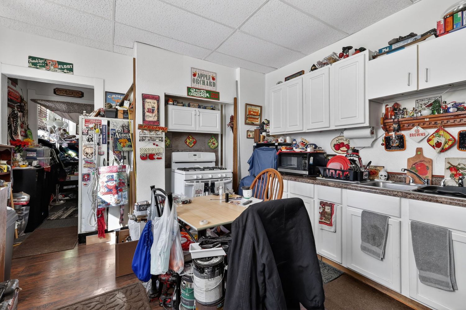 Cluttered kitchen with white cabinets, hardwood floors, drop ceiling, and vintage decor items covering countertops and walls.