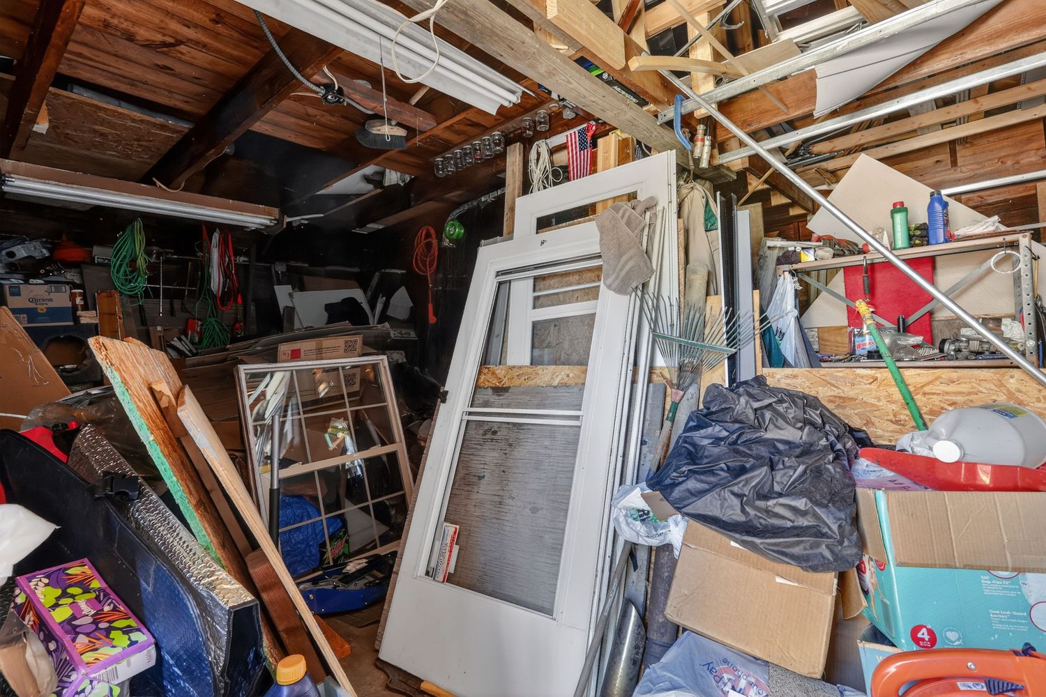 Cluttered garage storage area filled with old window frames, lumber, and miscellaneous items stacked against wooden walls.