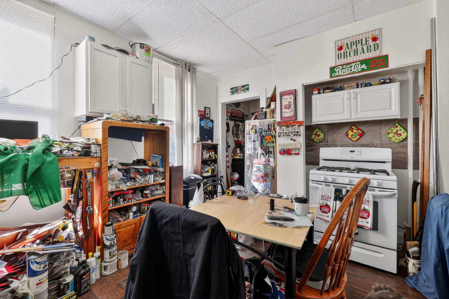 Cluttered kitchen with wooden dining table, white gas stove, and shelving units filled with various items.