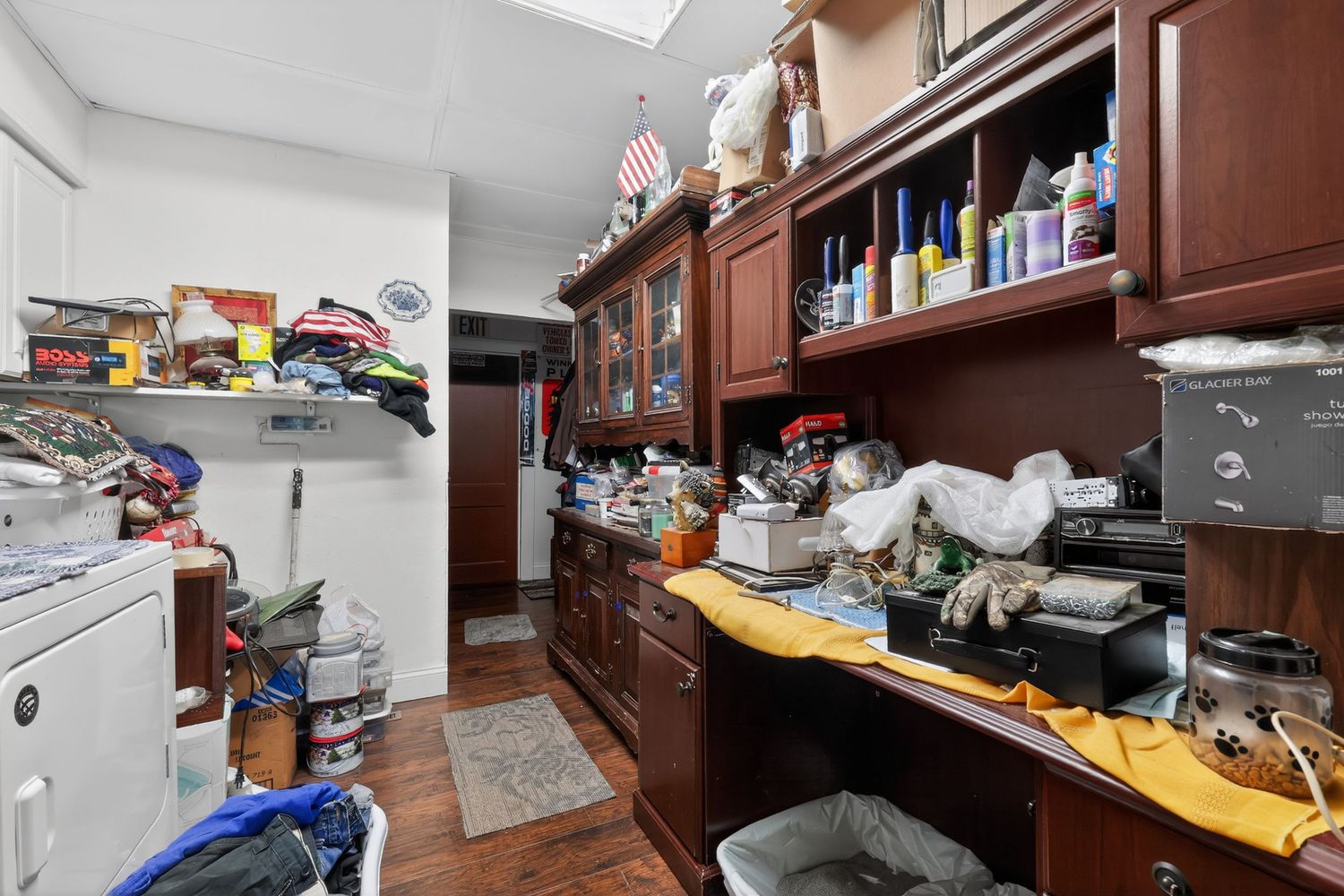 Cluttered utility room with dark wood built-in shelving, countertop workspace, and washer unit on hardwood floors.