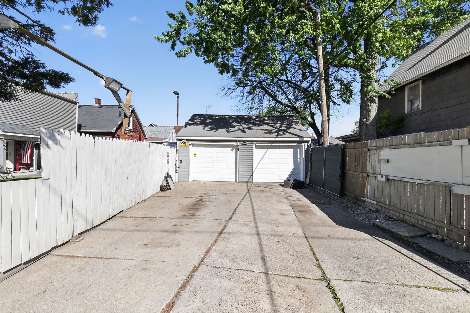 Spacious concrete driveway leading to two white garage doors, flanked by wooden fences and mature trees.