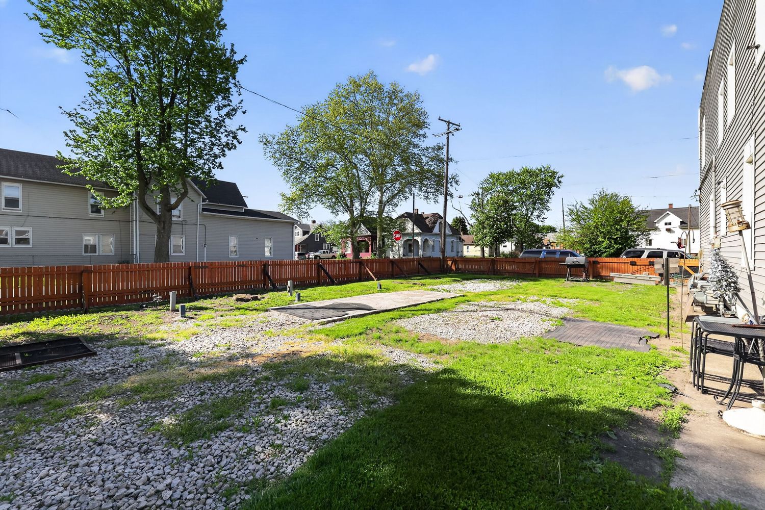 Vacant lot with gravel and grass next to a wooden fence, surrounded by residential homes and mature trees on a sunny day.