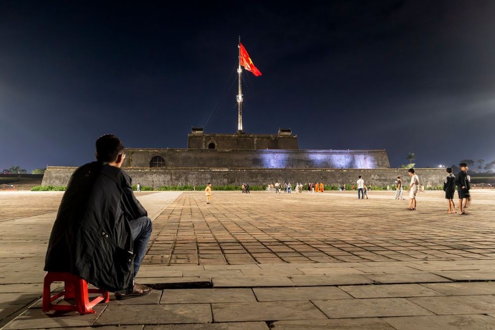 Person sitting at night near a flagpole with Vietnamese flag at Hue Citadel, Vietnam.