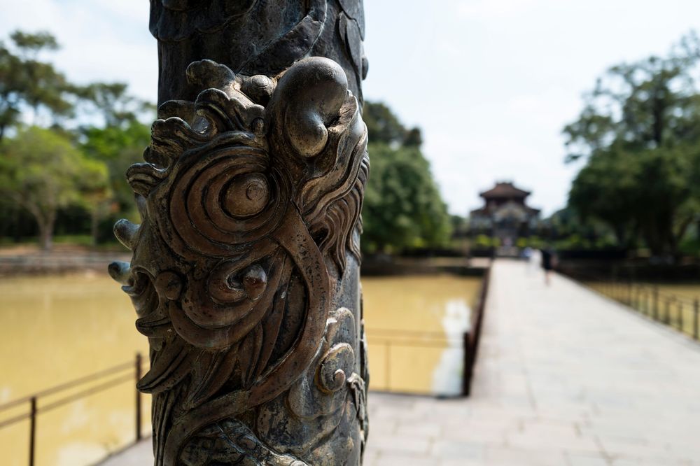 Ornate dragon-carved stone pillar at a Vietnamese temple with a serene pond and pagoda in the background.