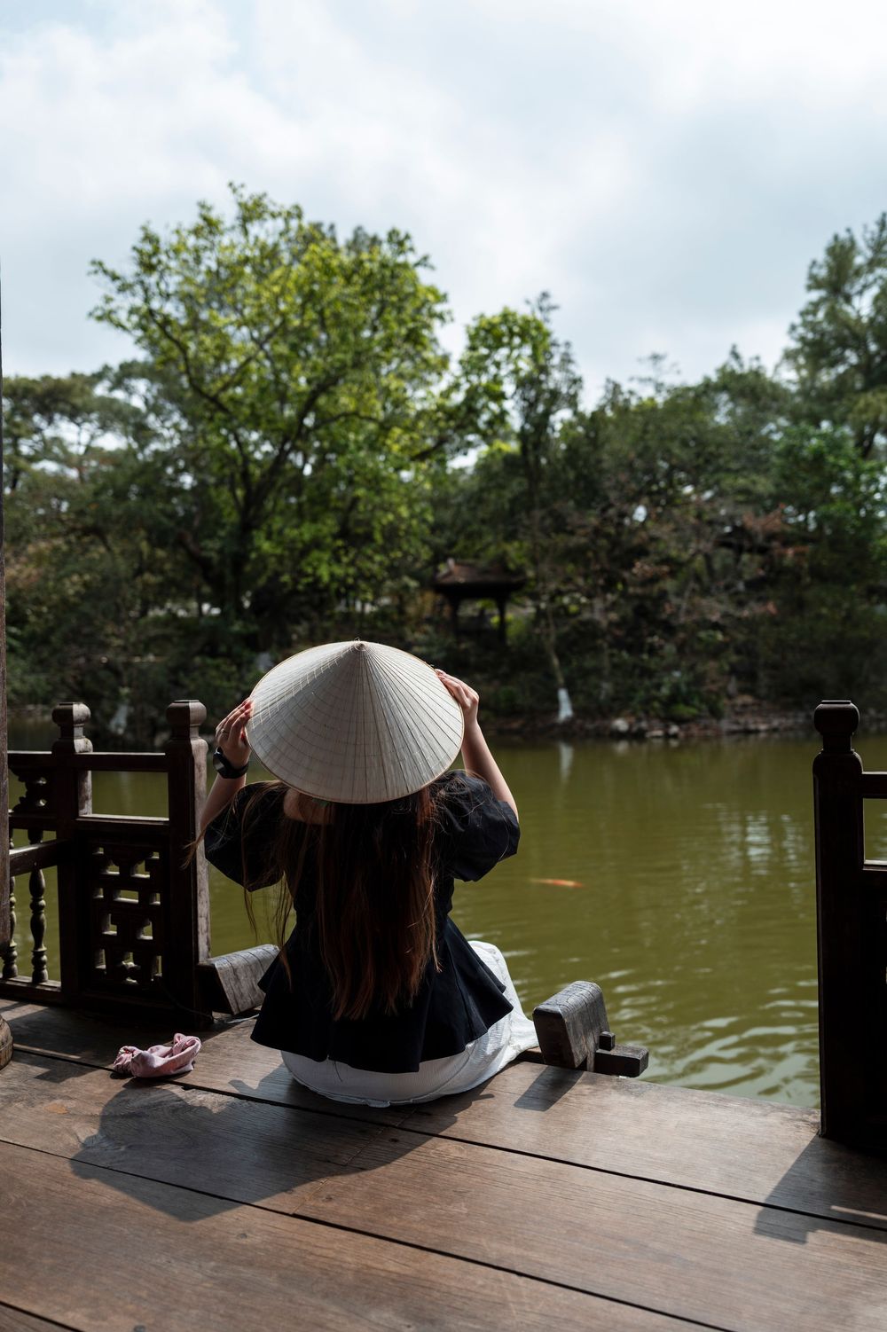 Woman in traditional Vietnamese conical hat sitting on wooden deck overlooking serene pond surrounded by lush green trees.