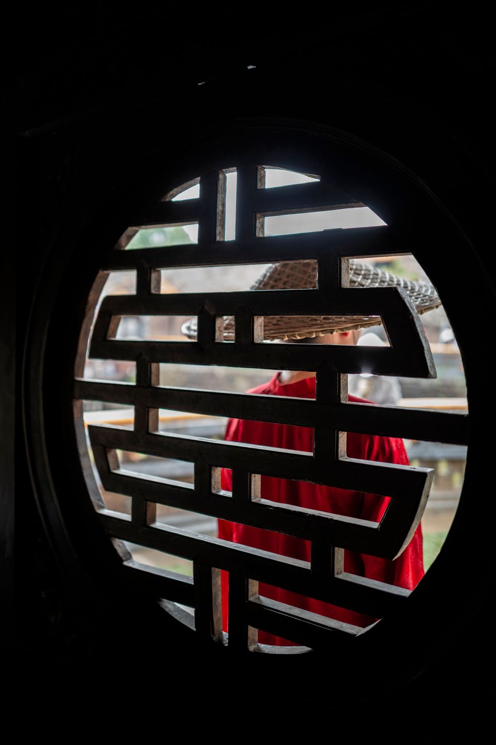 Person in red traditional Chinese attire viewed through ornate circular wooden lattice window pattern.