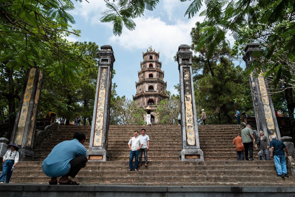Tourists climbing stone steps toward Thien Mu Pagoda tower in Hue, Vietnam, surrounded by lush trees.