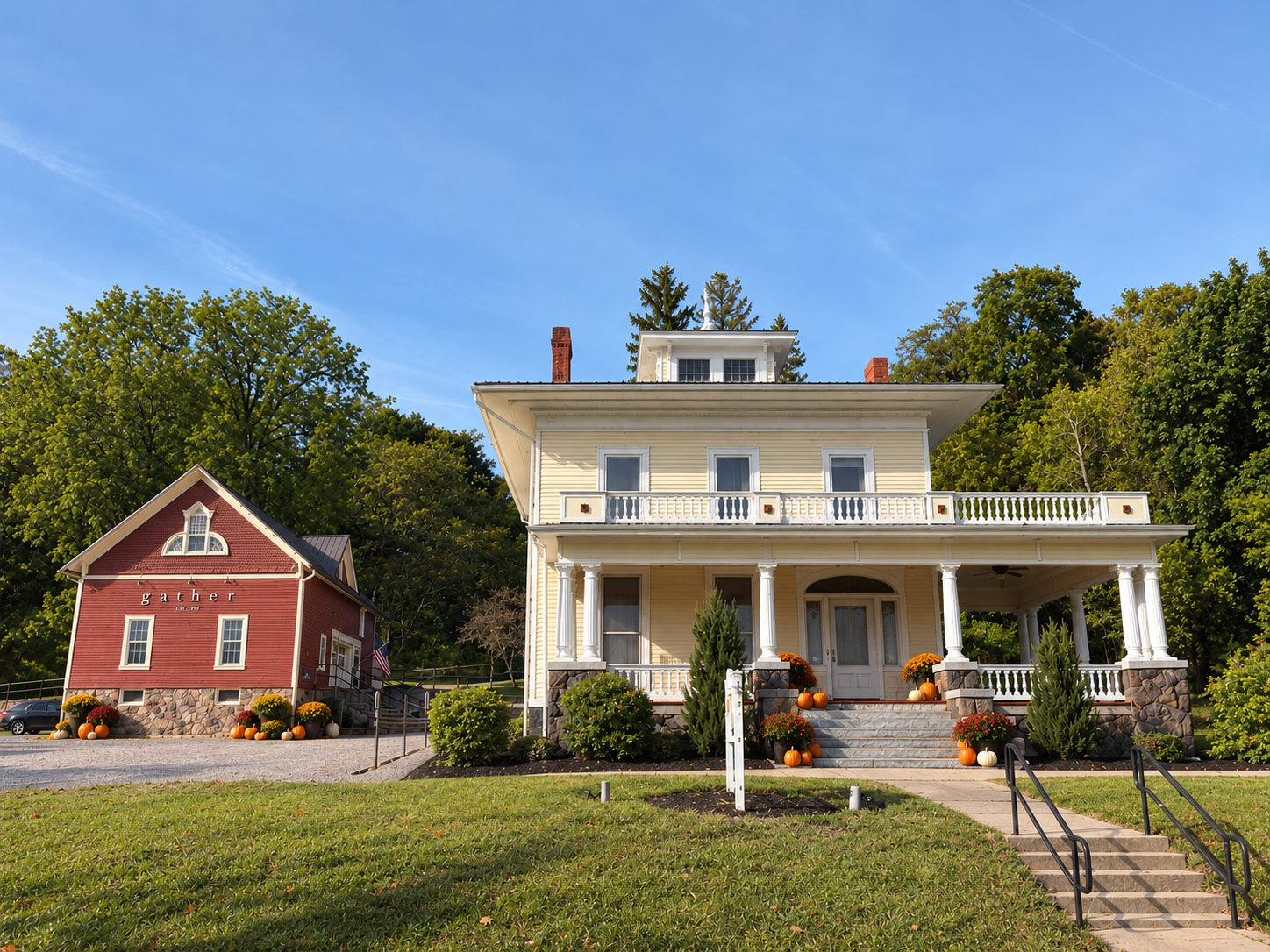 Large white colonial mansion with wrap-around porch beside a red brick building, surrounded by green trees and manicured lawn.