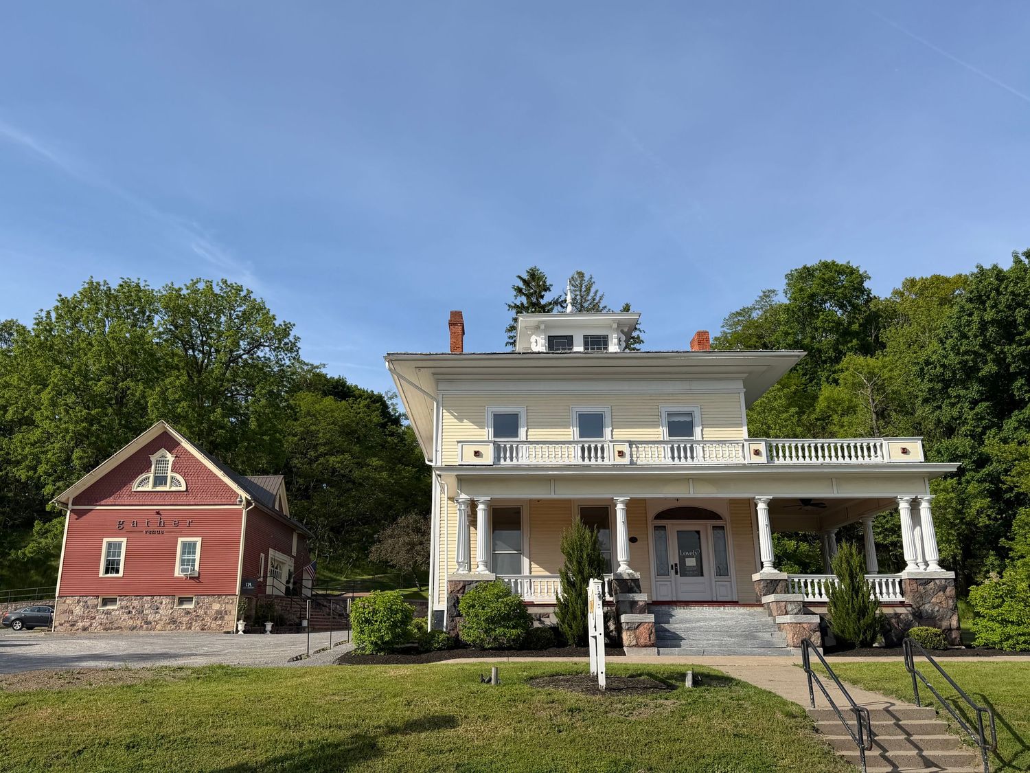 Large white colonial mansion with wrap-around porch beside a red brick building, surrounded by green trees and manicured lawn.