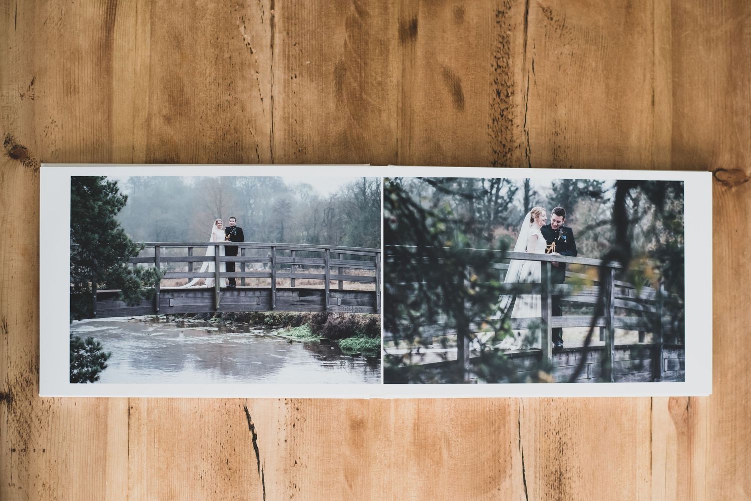 Open wedding photo album on wood surface showing bride and groom on a bridge in misty outdoor settings.