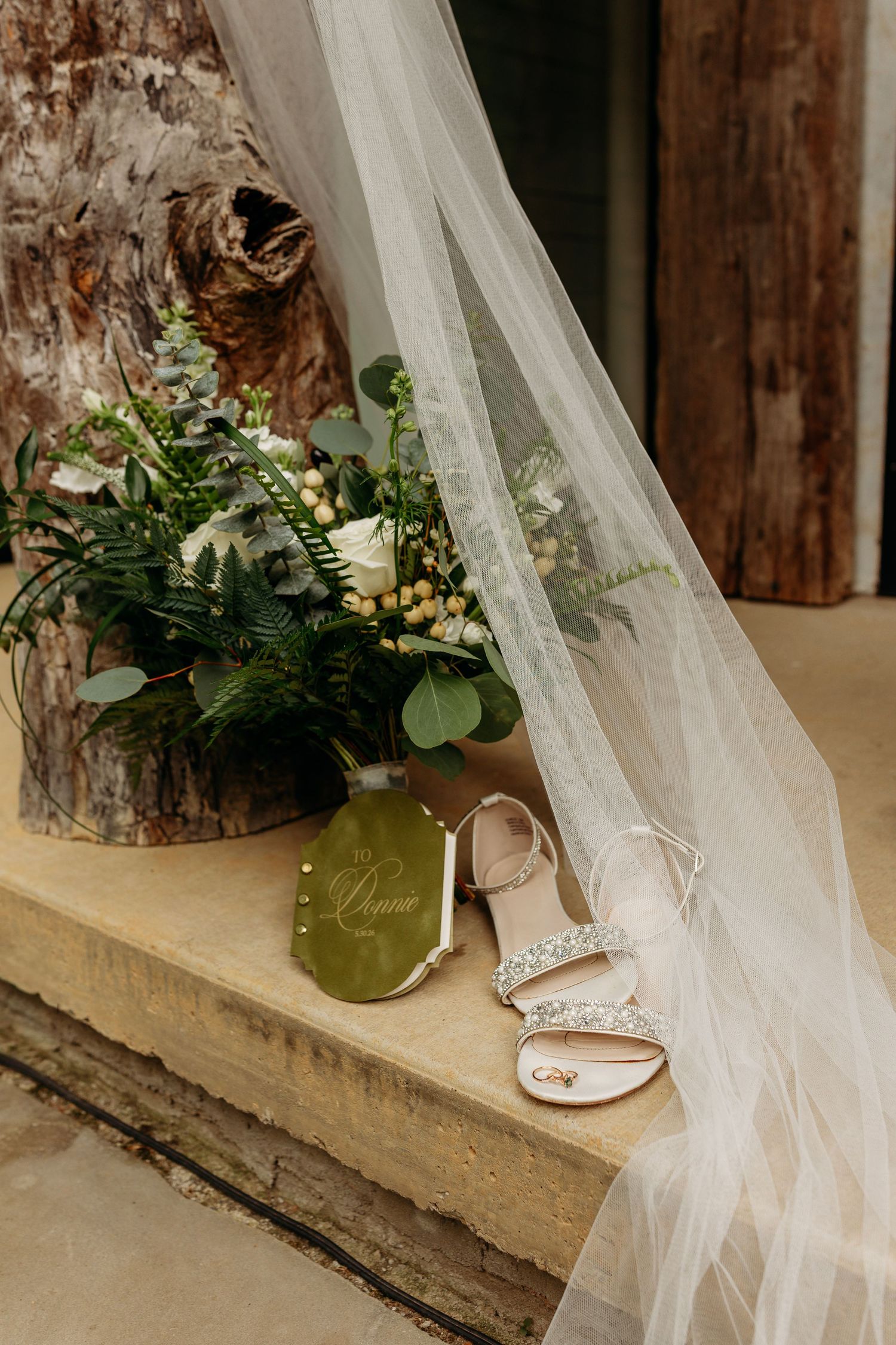 Bridal flat lay featuring crystal heeled sandals, lush greenery bouquet, veil, and olive green card on stone ledge.