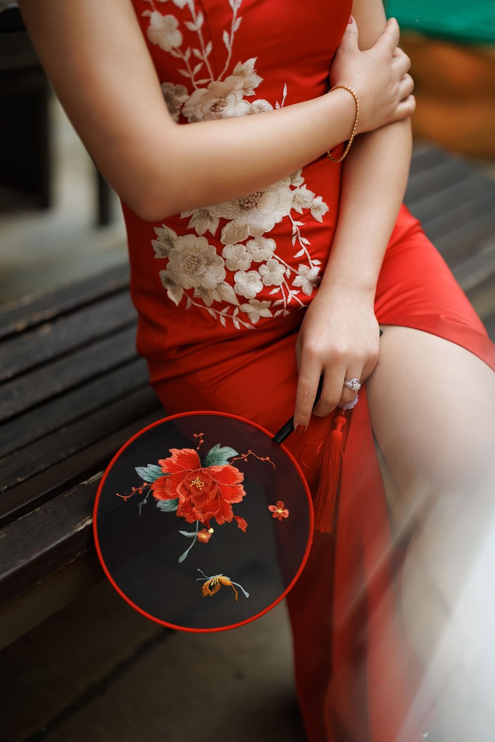 Close-up of red qipao dress with white floral embroidery, woman holding decorative Chinese fan with peony design.