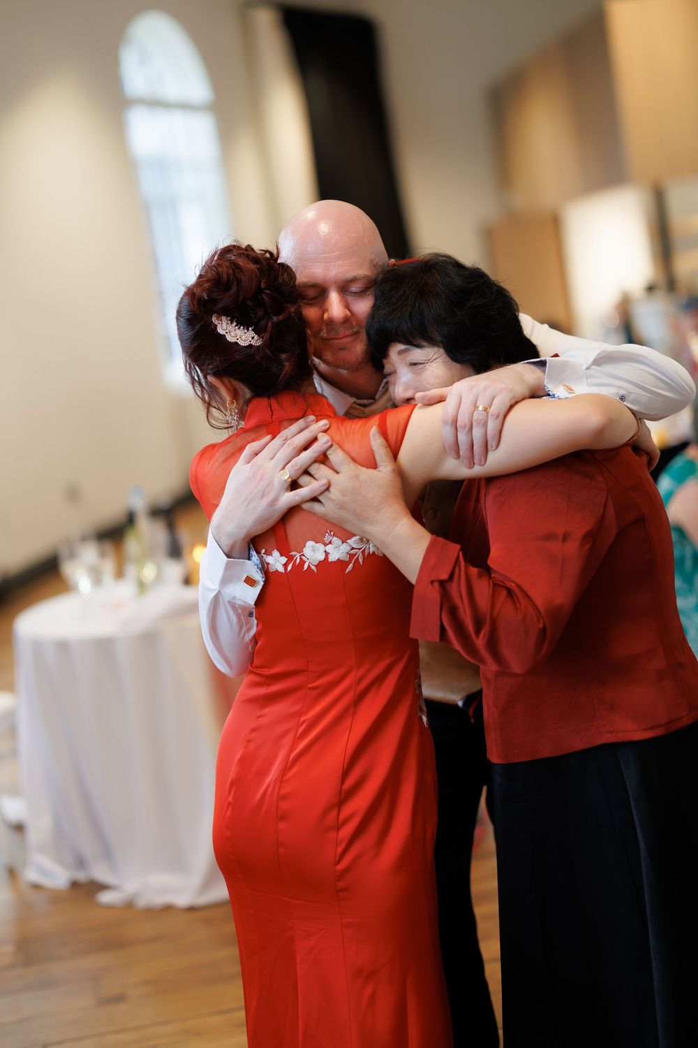A bald man in a suit embraces two women in red dresses at a wedding reception celebration.